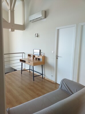 Quarto moderno e minimalista com uma escrivaninha de madeira, um sofá cinzento e um aparelho de ar condicionado fixado na parede.