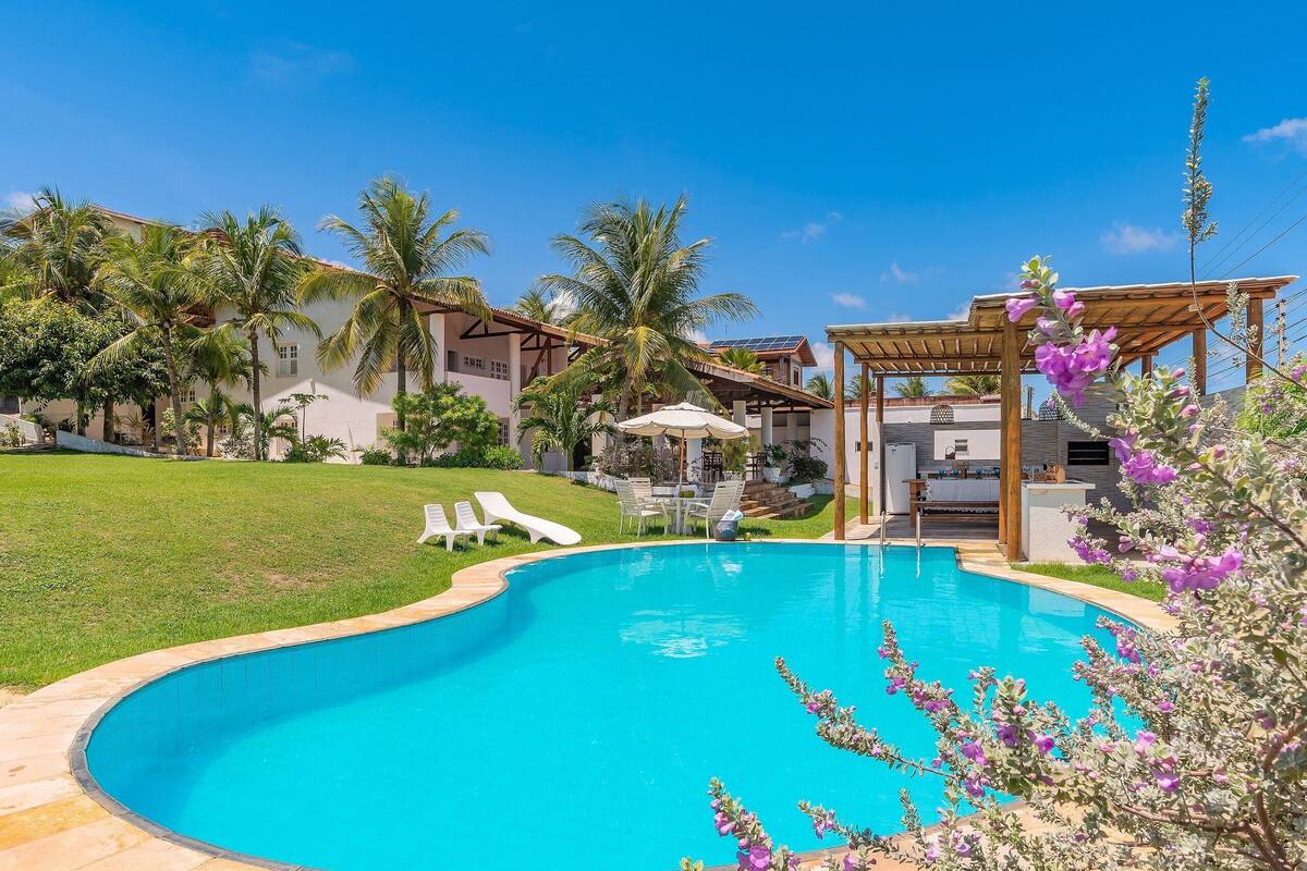 A swimming pool with a wooden deck, surrounded by palm trees and a covered patio area.