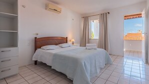 Ein Schlafzimmer mit Bett, Nachttisch, Kleiderschrank, einem Fenster mit Vorhängen und einer Klimaanlage.
