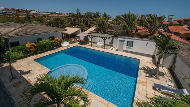 A rectangular swimming pool with a tiled deck, surrounded by palm trees and a small gazebo. - Casa Com Piscina em Porto das Dunas by Carpediem (Aquiraz)