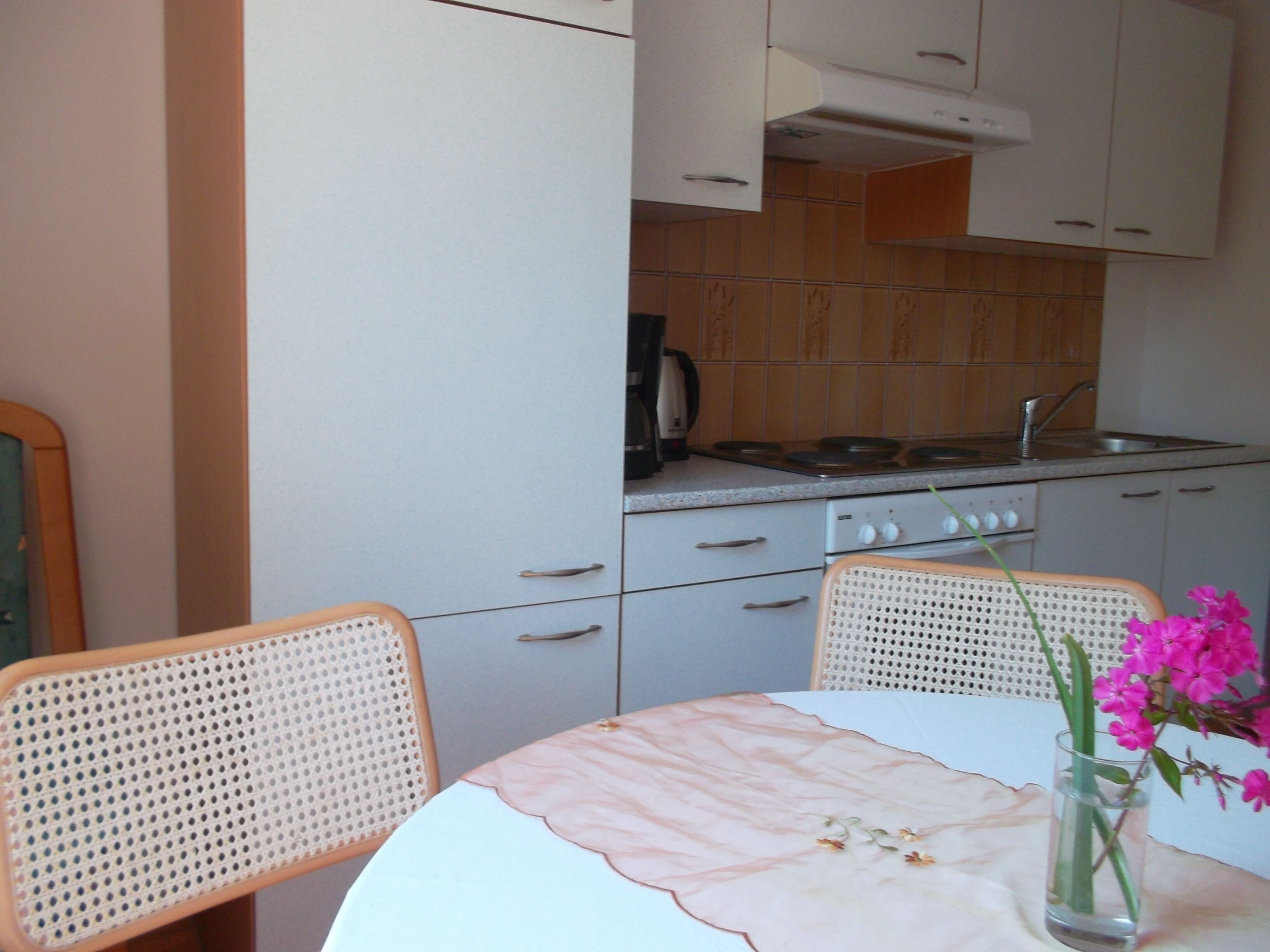 A kitchen with white cabinets, a stove, and a sink. There is a table with a vase of flowers and two wicker chairs.