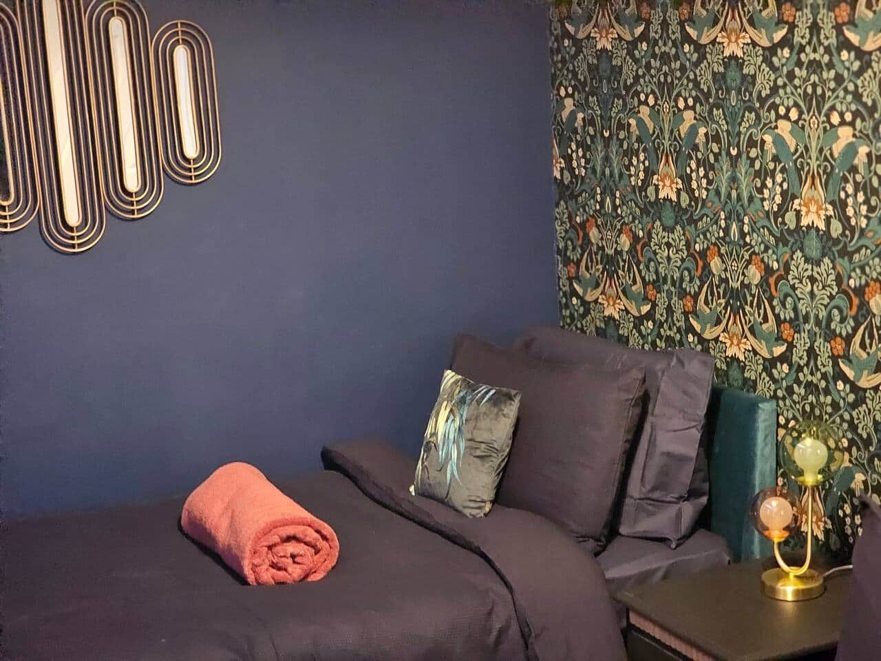 Een slaapkamer met een donkerblauwe wand, behang met een patroon, een bed met donkere beddengoed, een opgerolde handdoek en een nachtkastje met een lamp.