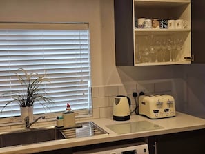 Une cuisine avec une fenêtre, un évier, une bouilloire, un grille-pain et une étagère où sont rangées des tasses et des verres.