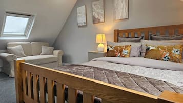 A bedroom with a sloped ceiling, a bed with a wooden headboard, a nightstand, a lamp, a sofa, and a window.