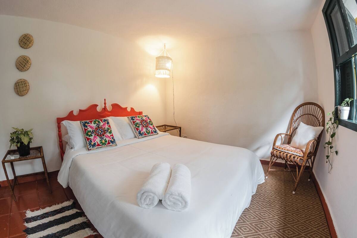 Ein ordentlich gemachtes Bett mit weißen Leinentüchern und zwei aufgerollten Handtüchern, ein Korbstuhl mit Kissen, ein kleiner Tisch mit Pflanze und eine Wandlampe.