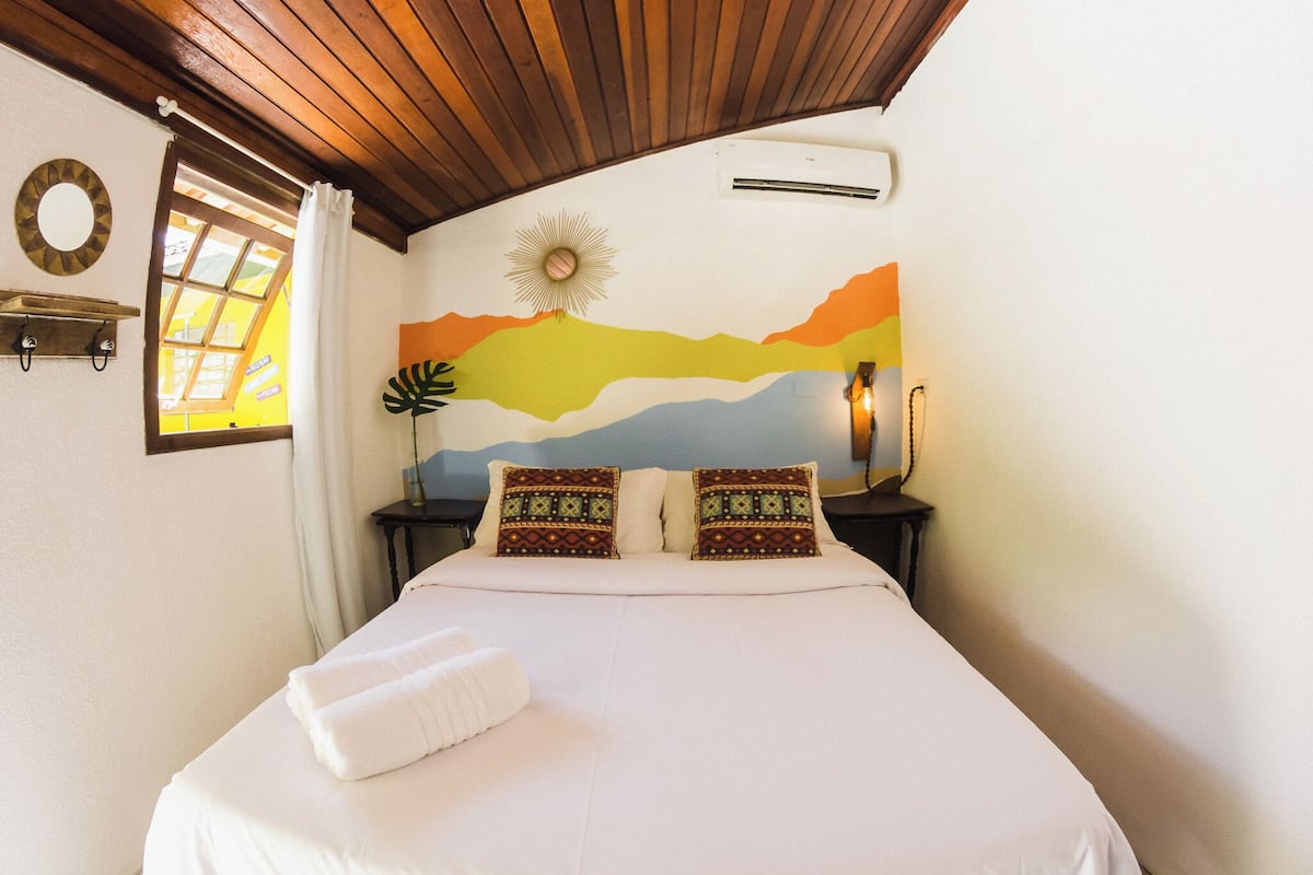 Ein Schlafzimmer mit einem Bett, einem Nachttisch, einer Wand-Klimaanlage, einem Fenster mit Vorhängen und einem Wanddekor.