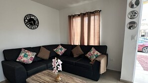 Uma sala de estar com um sofá preto, uma mesa de centro de madeira, um relógio de parede e uma janela com cortinas.