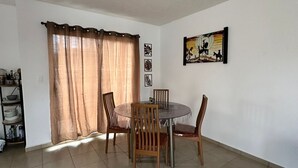 Uma área de refeições com uma mesa redonda e quatro cadeiras, uma janela com cortinas, um candeeiro fixado na parede e um quadro emoldurado.