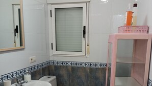 Une salle de bain avec un lavabo, des toilettes et un miroir. Il y a une fenêtre avec un panneau en verre dépoli et une étagère rose avec des rangements.