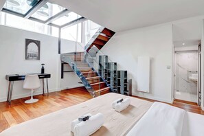 Une chambre d’hôtel moderne avec un plancher en bois, un lit recouvert de draps blancs, un bureau avec une chaise, un escalier en verre et une vue sur la ville par la fenêtre.