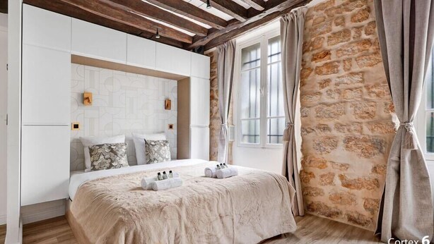 Un dormitorio con pared de piedra, techo de madera y cama con manta y almohadas color beige.