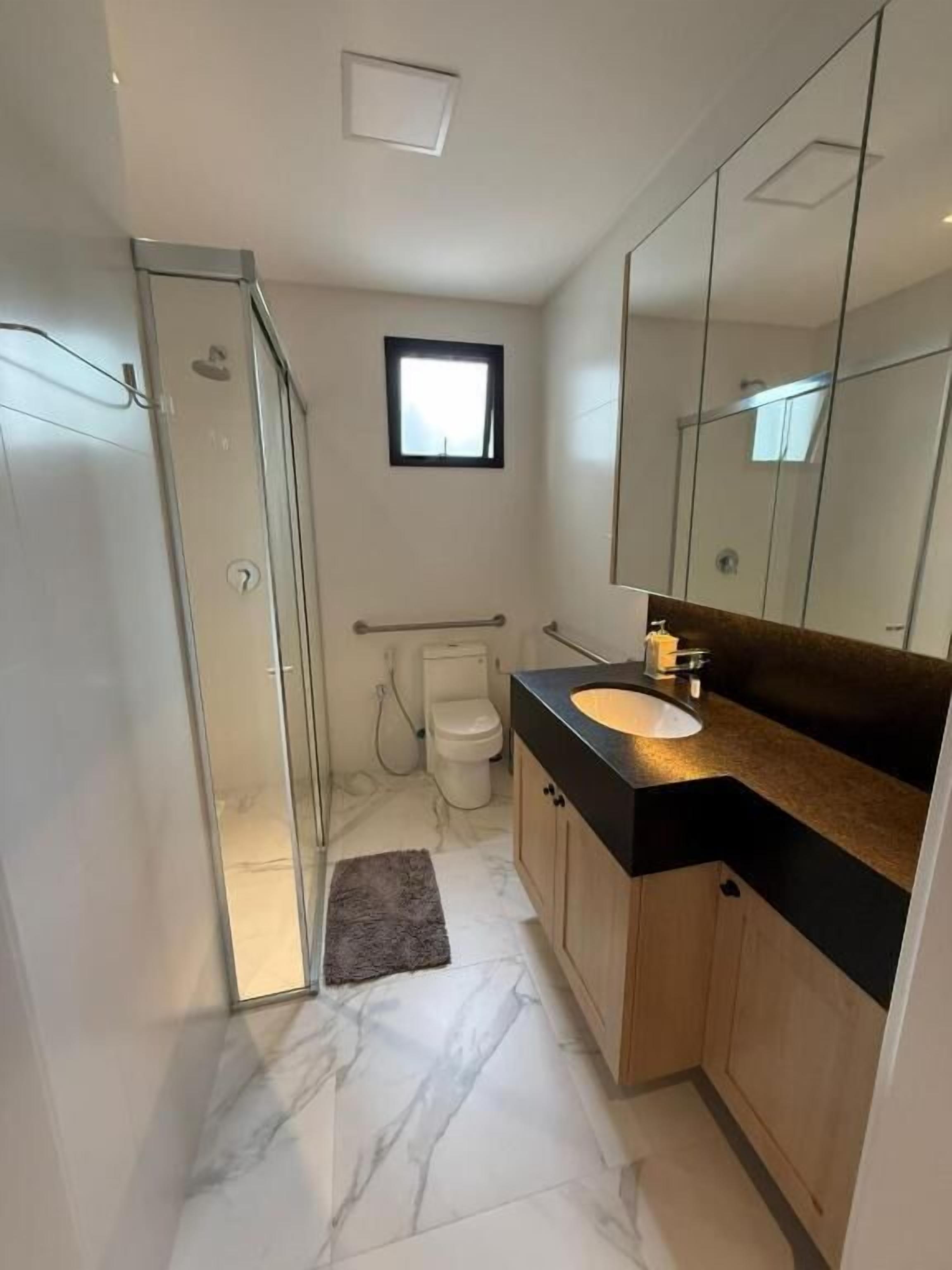 Un baño moderno con cabina de ducha de vidrio, inodoro, lavabo con espejo y una ventana pequeña.
