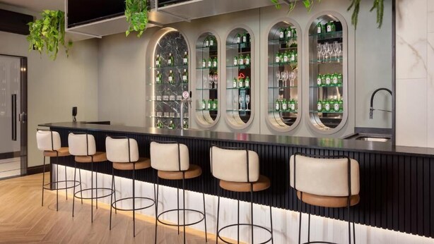 Un área de bar moderna con una barra larga, varios taburetes y estantes llenos de botellas y copas.
