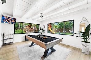 A modern indoor pool table with a wooden frame and black felt, surrounded by large windows offering views of a backyard with a slide and play equipment. - Luxury 4BR Retreat w/ Heated Pool, Jacuzzi® Hot Tub & Indoor/Outdoor Games (Hollywood)