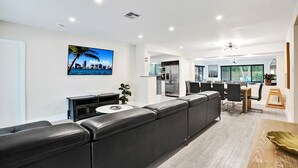 Una sala moderna con un sofá de cuero negro, un comedor con una mesa de madera y una cocina al fondo.