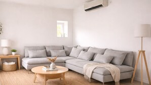 Uma sala de estar moderna com um sofá seccional cinza, uma mesa de centro redonda de madeira, um abajur e um ar condicionado fixado na parede.