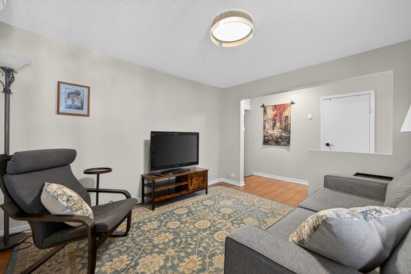 A living room with a flat-screen TV on a wooden stand, grey sofas, a patterned rug, and a painting on the wall.