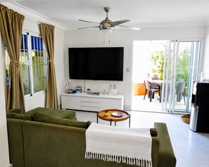 Ein Wohnzimmer mit einer Couch, einem Couchtisch, einem Flachbildfernseher, einem Deckenventilator und einer Schiebetür zu einem Außenbereich mit Tisch und Stühlen.