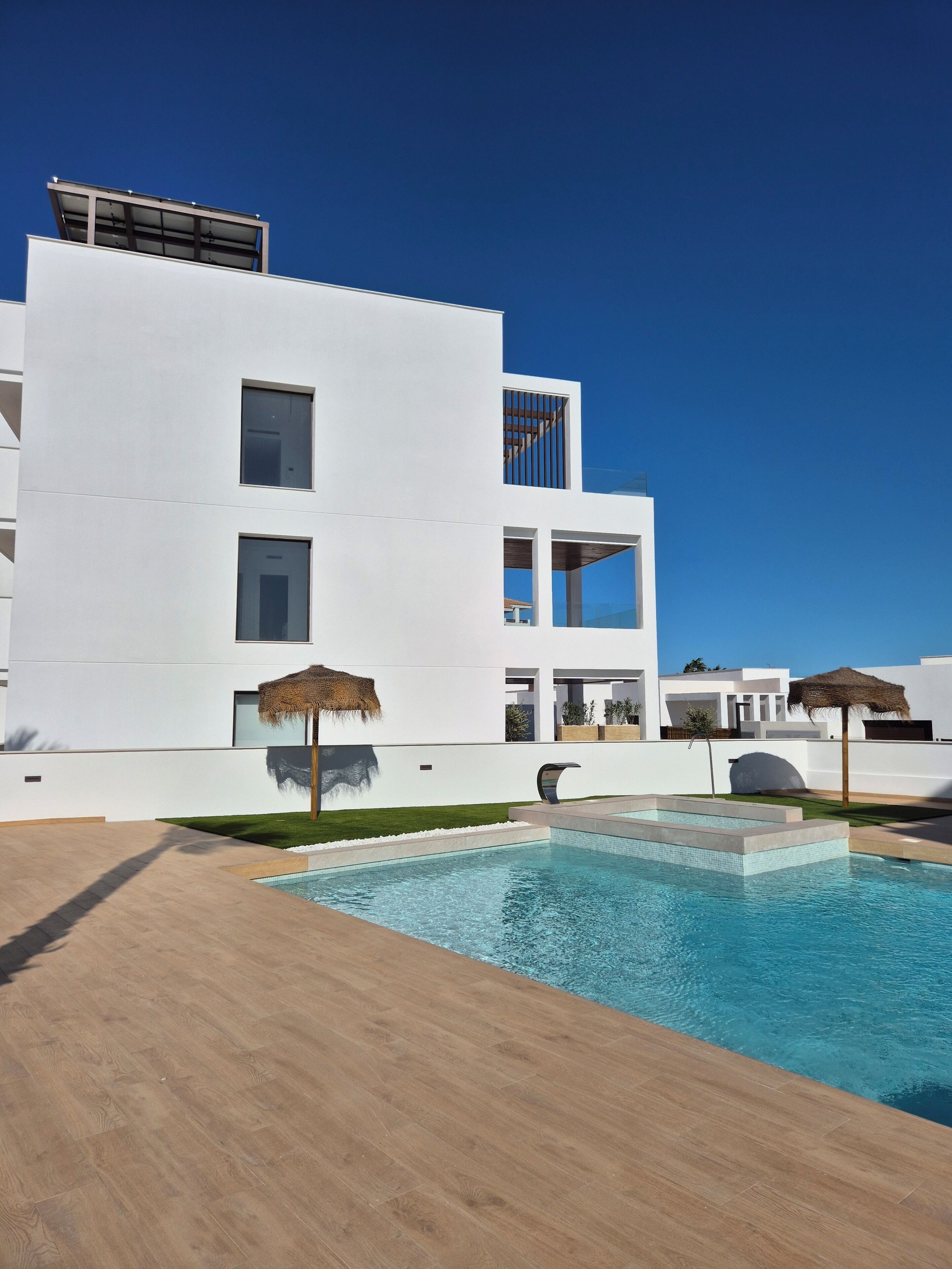 Un bâtiment moderne doté d’une piscine, d’une façade blanche et de parasols en chaume.