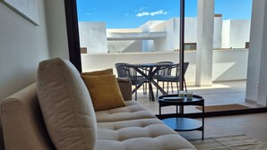 Una sala de estar moderna con un sofá color crema, una mesita de centro y una zona de comedor con sillas y una mesa. Un gran ventanal corredero da a una terraza exterior con vistas al cielo y edificios.