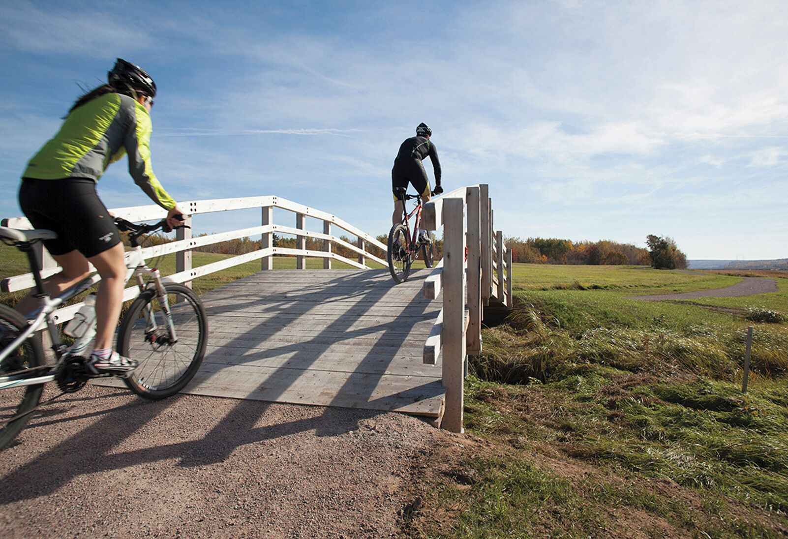 A wooden bridge with white railings, cyclists on it, and a grassy landscape in the background.