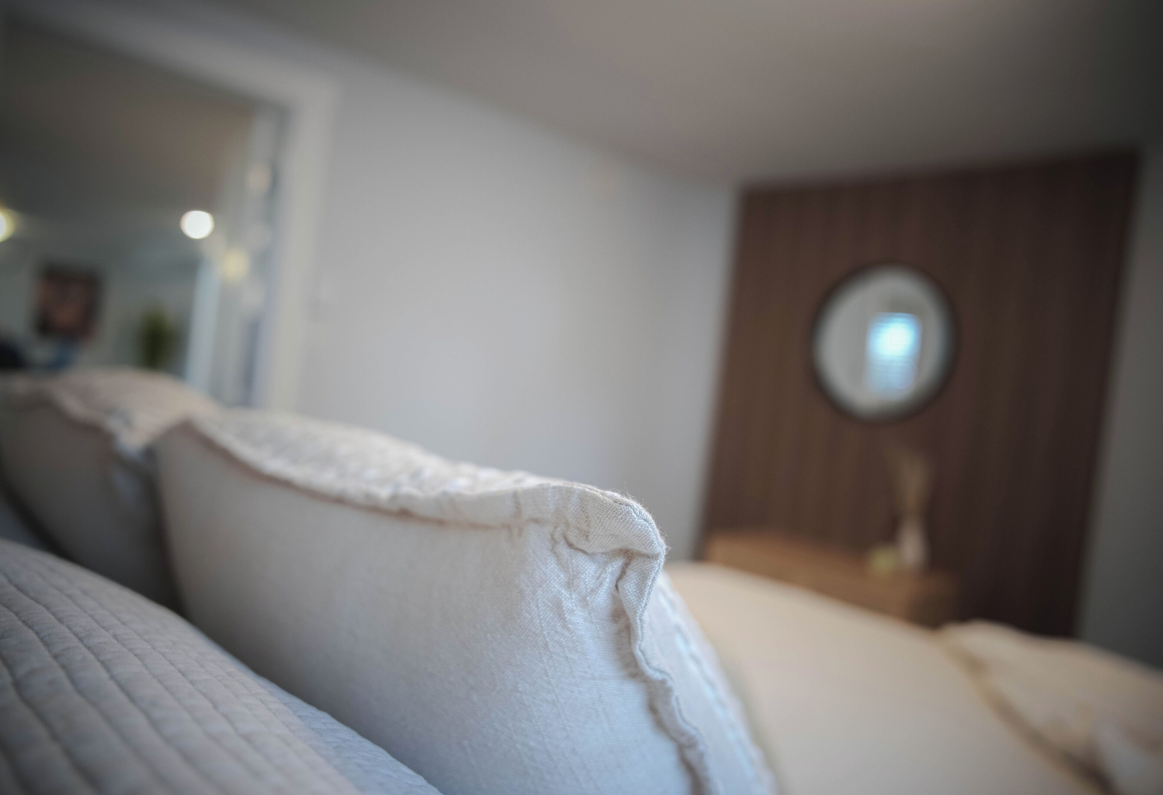 A close-up of a bed with white bedding, a wooden headboard, and a round mirror on a wooden nightstand.