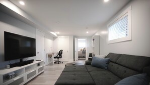 A modern living room with a large flat-screen TV, a grey sofa, and a white entertainment unit. - Warm and Welcoming Basement (Dieppe)