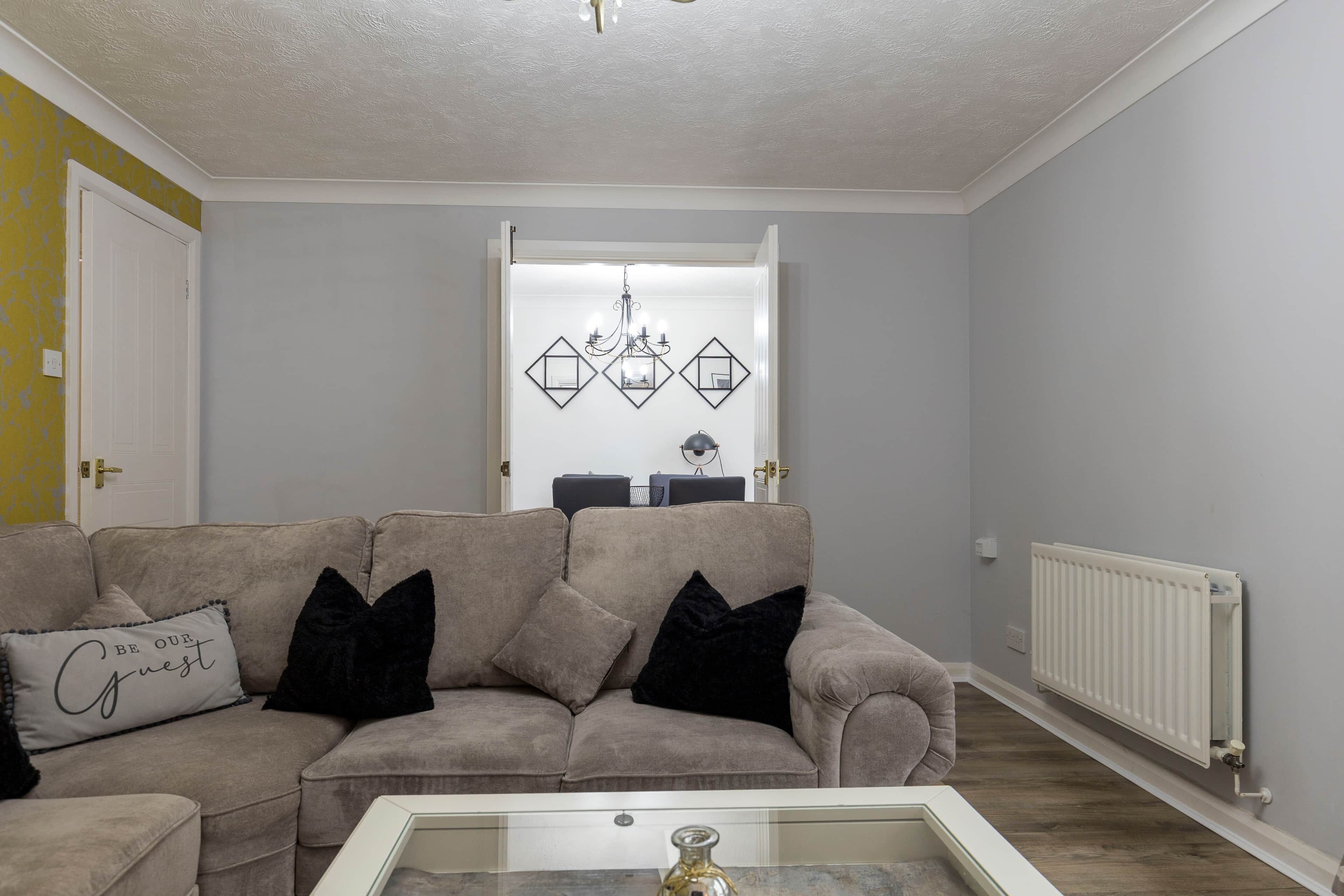 Een woonkamer met een beige bank, zwarte kussens, een glazen salontafel en een kroonluchter.