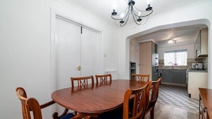 Uma sala de jantar com mesa e cadeiras de madeira, um candelabro e vista para a cozinha.