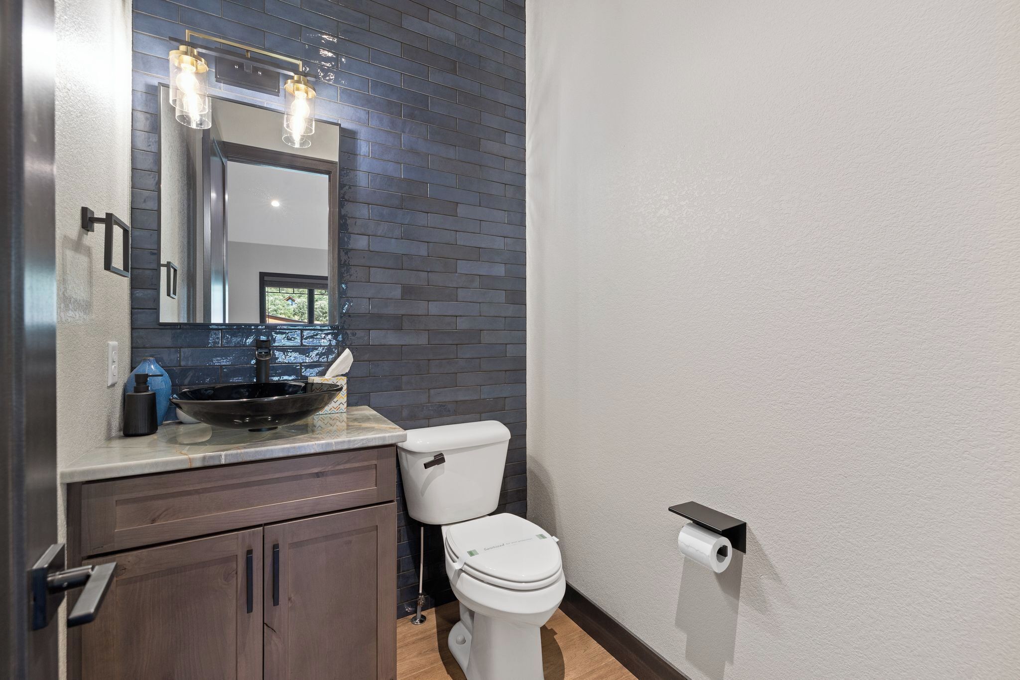A bathroom with a dark tiled wall, a white toilet, a sink with a bowl, and a mirror with lights.