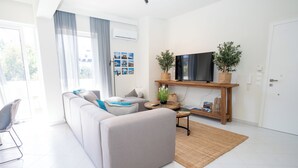 A modern living room with a grey sofa, a flat-screen TV on a wooden stand, and a large window with sheer curtains. - Design-Apartment mit 2 Balkonen & gehobener Ausstattung (Rodos)