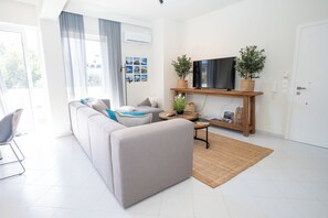 Ein modernes Wohnzimmer mit einem grauen Sofa, einem Flachbildfernseher auf einem Holzboden und einem großen Fenster mit durchscheinenden Vorhängen.