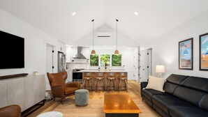 Ein modernes Wohnzimmer mit einer schwarzen Ledersofa, einem Holztisch und einem grauen Pouf. Im Hintergrund ist eine Küche mit weißen Schränken und einer zentralen Kochinsel mit Barhocker erkennbar.