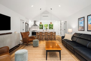 Ein modernes Wohnzimmer mit einer schwarzen Ledersofa, einem Holztisch und einem grauen Pouf. Im Hintergrund ist eine Küche mit weißen Schränken und einer zentralen Kochinsel mit Barhocker erkennbar.