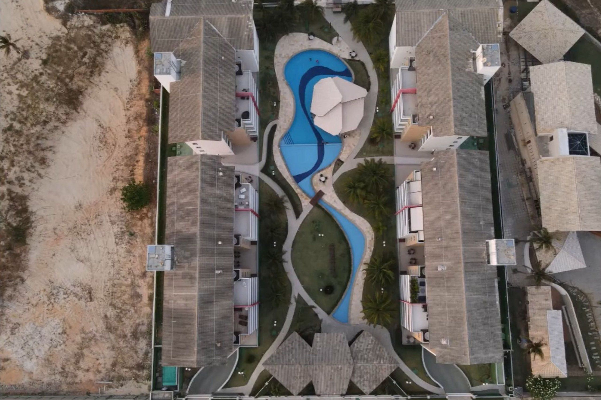 Vista aérea de un complejo turístico con una piscina central, varias edificaciones y una zona de playa arenosa.