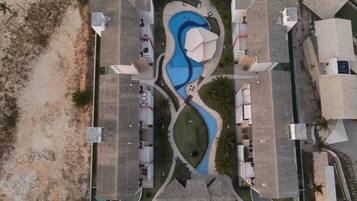 Vista aérea de un complejo turístico con una piscina central, varias edificaciones y una zona de playa arenosa.