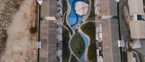 An aerial view of a resort with a central swimming pool, multiple buildings, and a sandy beach area.