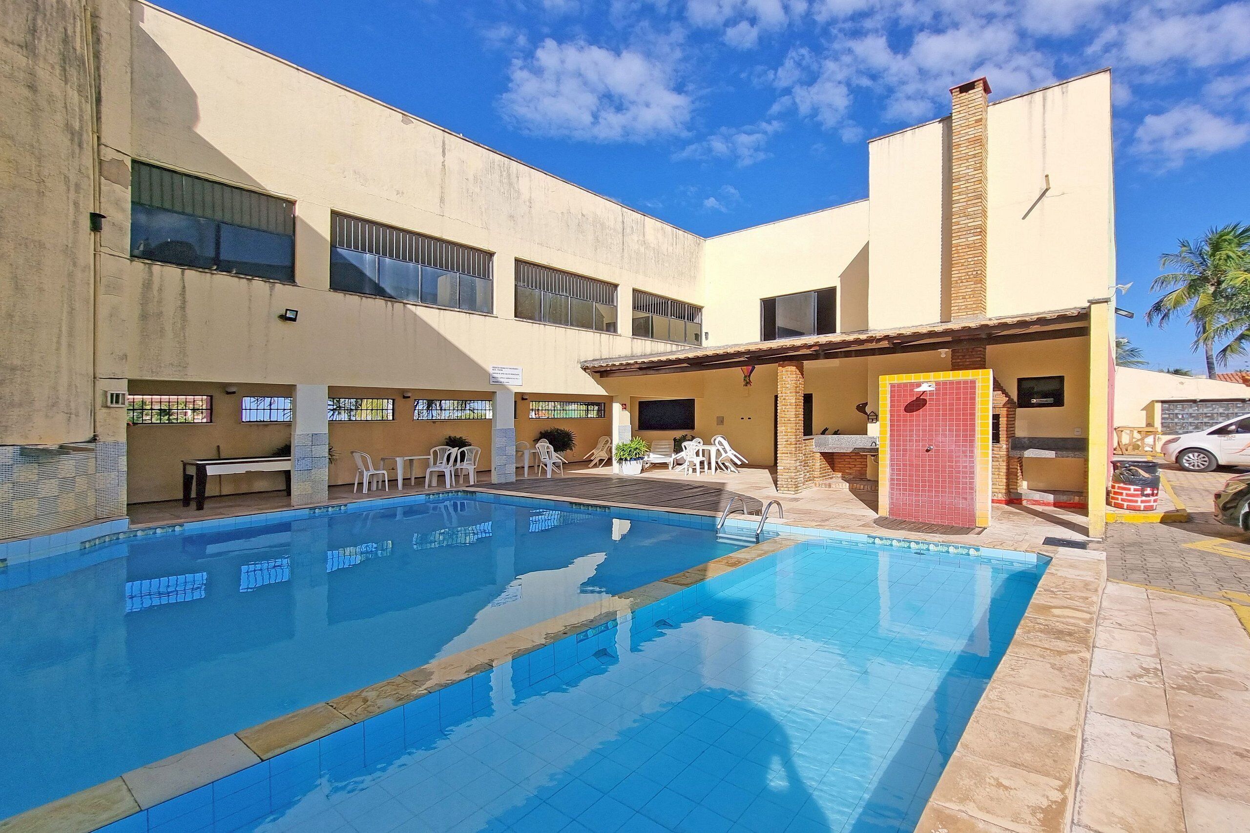Una piscina con bordes de azulejos, rodeada de mobiliario exterior y un edificio con varias ventanas y una puerta roja.