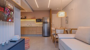 A modern kitchen with a refrigerator, microwave, and wooden cabinets. There is a dining area with a table and chairs, and a sofa in the living room. - Crystal Park #206 - Quadruplo por Carpediem (Aquiraz)
