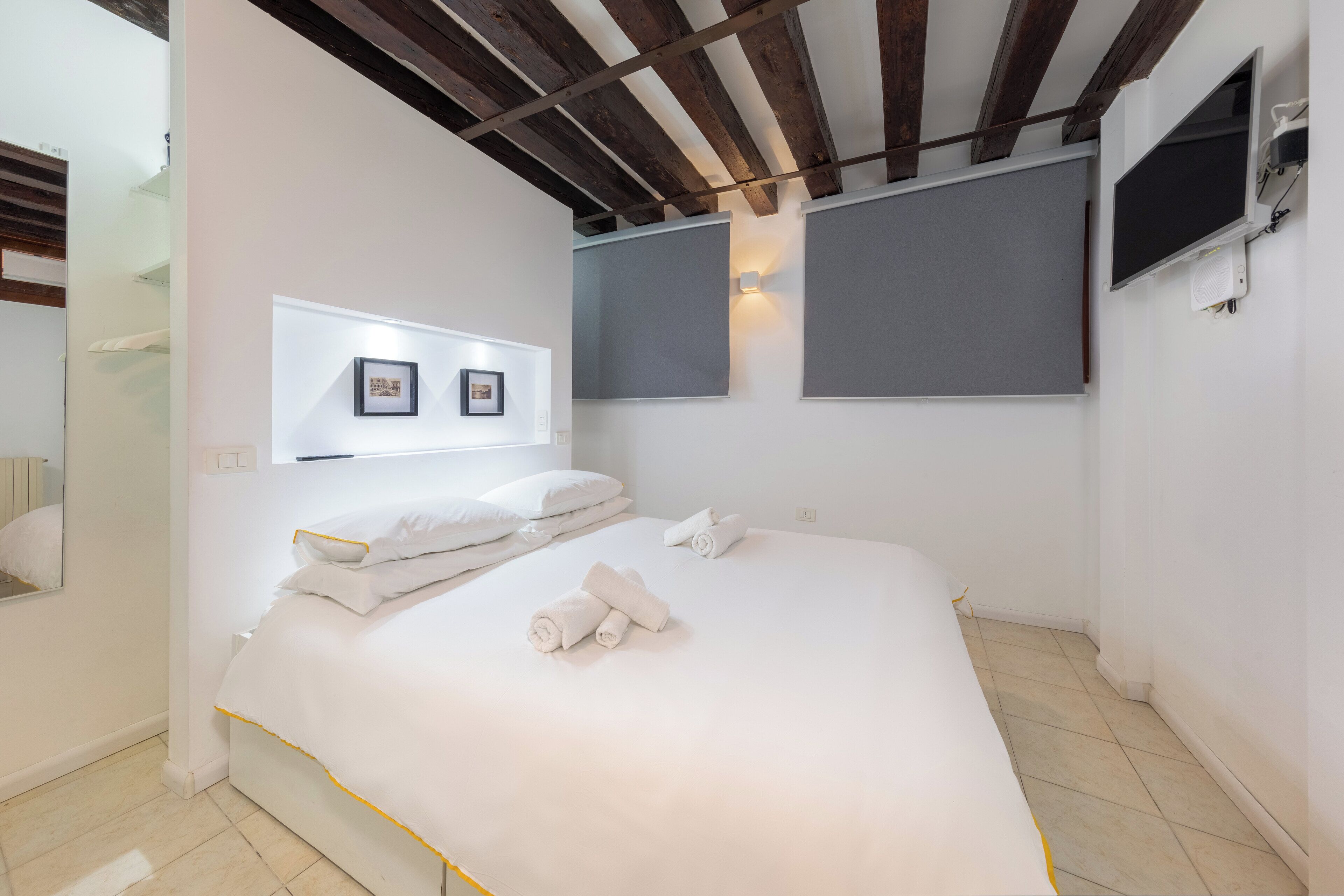 A modern hotel room with a large bed, white linens, a wooden ceiling, and a flat-screen TV mounted on the wall.