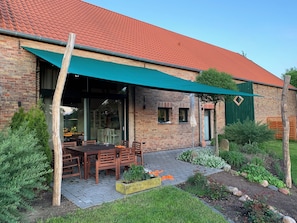 A brick building with a red-tiled roof, a covered patio with wooden furniture, and a garden with various plants and a small pond. - Old barn in the Nemitzer Heide (Trebel)
