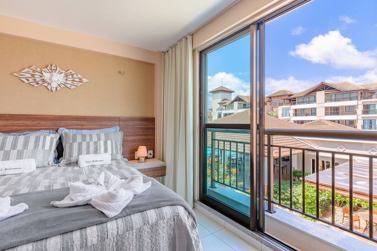 A hotel room with a large window, a bed with labeled pillows, and a decorative wall piece.