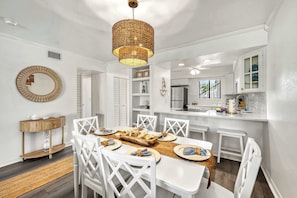 A dining area with a white table set for six, white chairs, a woven centerpiece, and a hanging light fixture. - Newly Rehabbed • Pickleball/Tennis • Beach Access (Bradenton Beach)