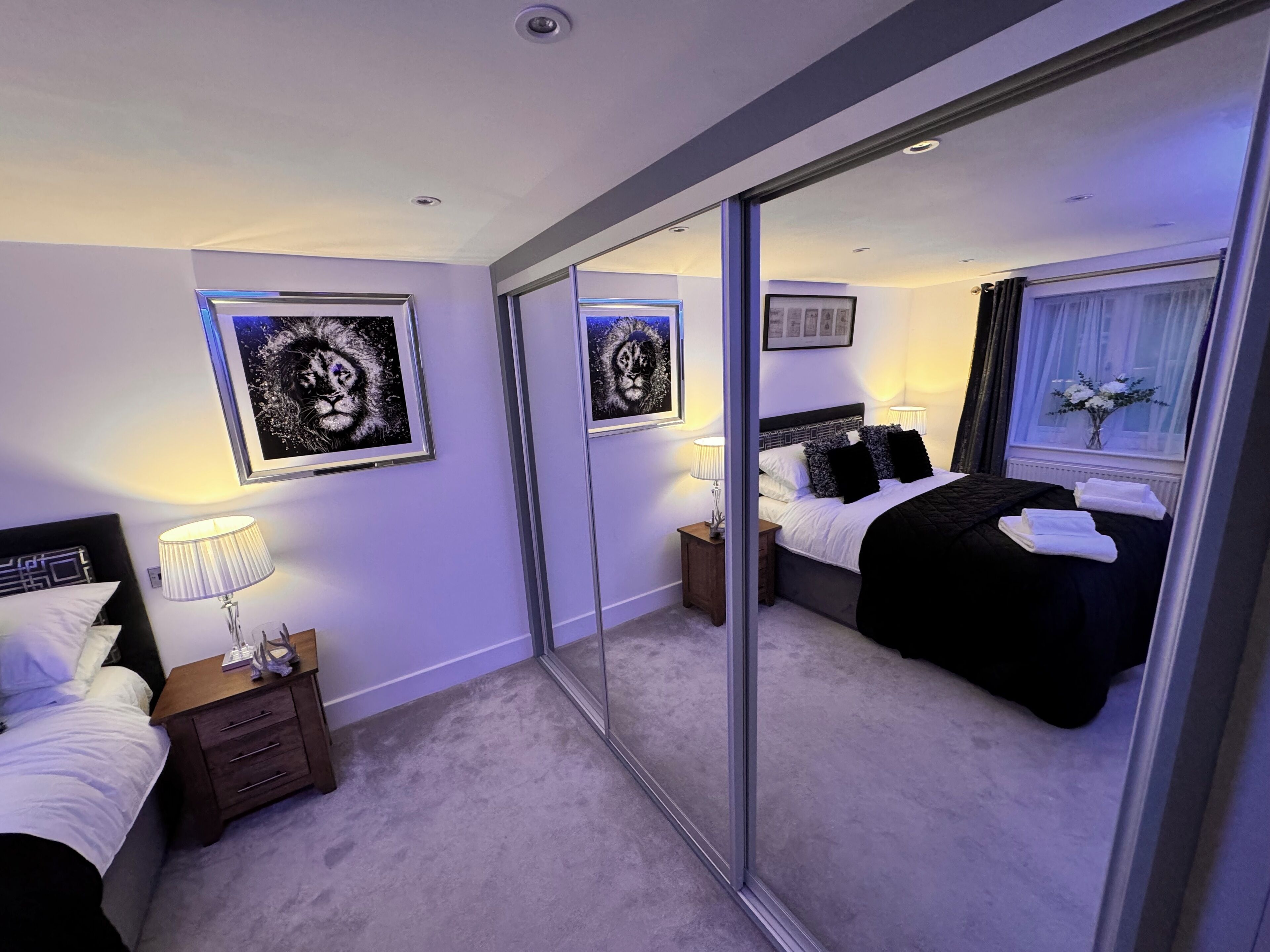 A modern hotel room with a large mirror, a neatly made bed, a bedside table with a lamp, and framed artwork on the wall.