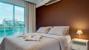 A hotel room with a large bed, a nightstand, a lamp, and a view of buildings through the window. - Palm Beach #201 - Porto das Dunas por Carpediem (Aquiraz)