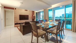 A modern dining area with a glass table, wooden chairs, and a flat-screen TV mounted on the wall. - Palm Beach #201 - Porto das Dunas por Carpediem (Aquiraz)