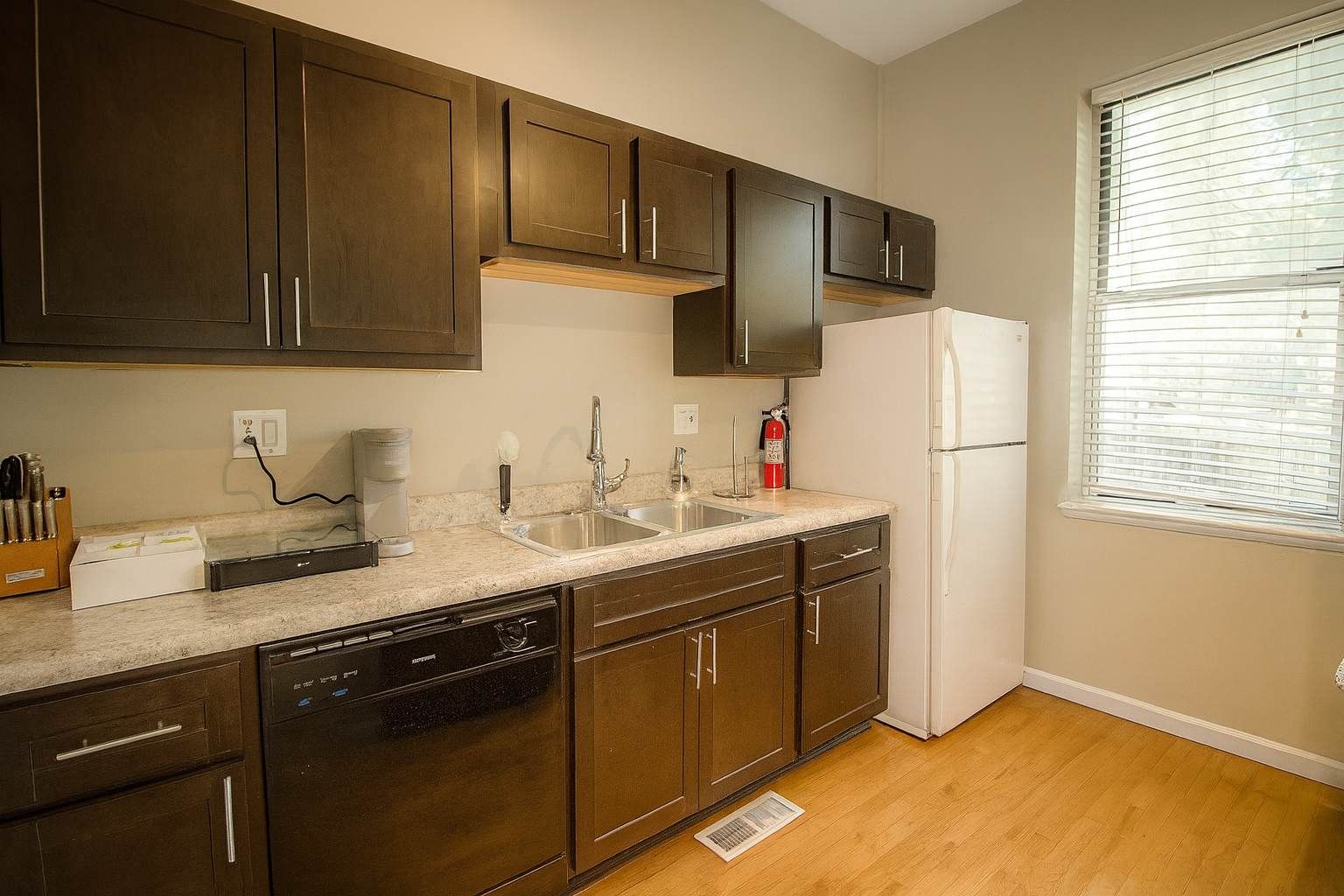 A kitchen with dark wooden cabinets, a white refrigerator, and a window with blinds.