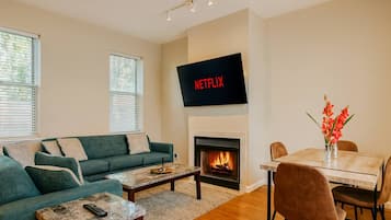 A living room with a green sofa, a coffee table, a fireplace, a dining table with chairs, and a TV mounted above the fireplace.