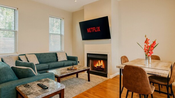 A living room with a green sofa, a coffee table, a fireplace, a dining table with chairs, and a TV mounted above the fireplace.