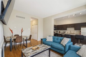 Una sala de estar moderna con un sofá azul, una mesa de comedor con sillas, y una cocina con armarios oscuros y un refrigerador blanco.
